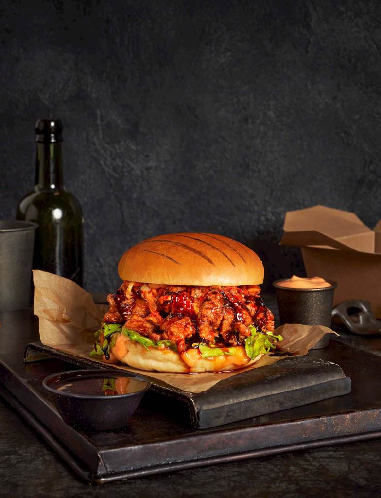 A display focused on an Americana Grill Marked Vegan Brioche Style Bun filled with soy glazed fried chicken, korean bbq sauce, and kimchi.