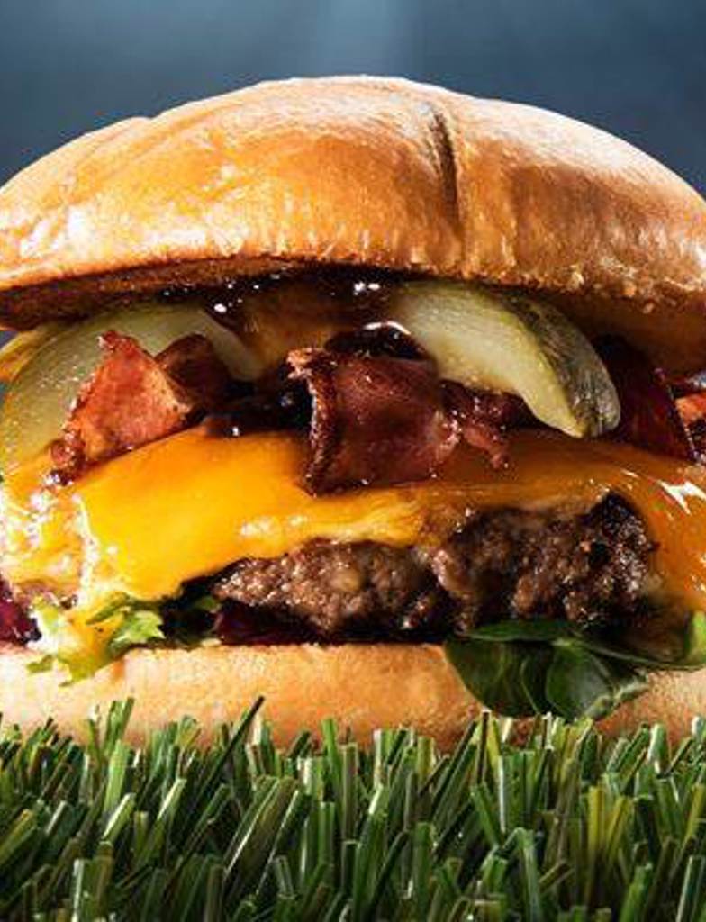 a massive burger is on display in a football stadium looking impressive on the astroturf, the burger is compiled of an Americana brioche bun, salad greens, beef burger patty, melted cheddar cheese, bacon, pickle spear, and bbq sauce