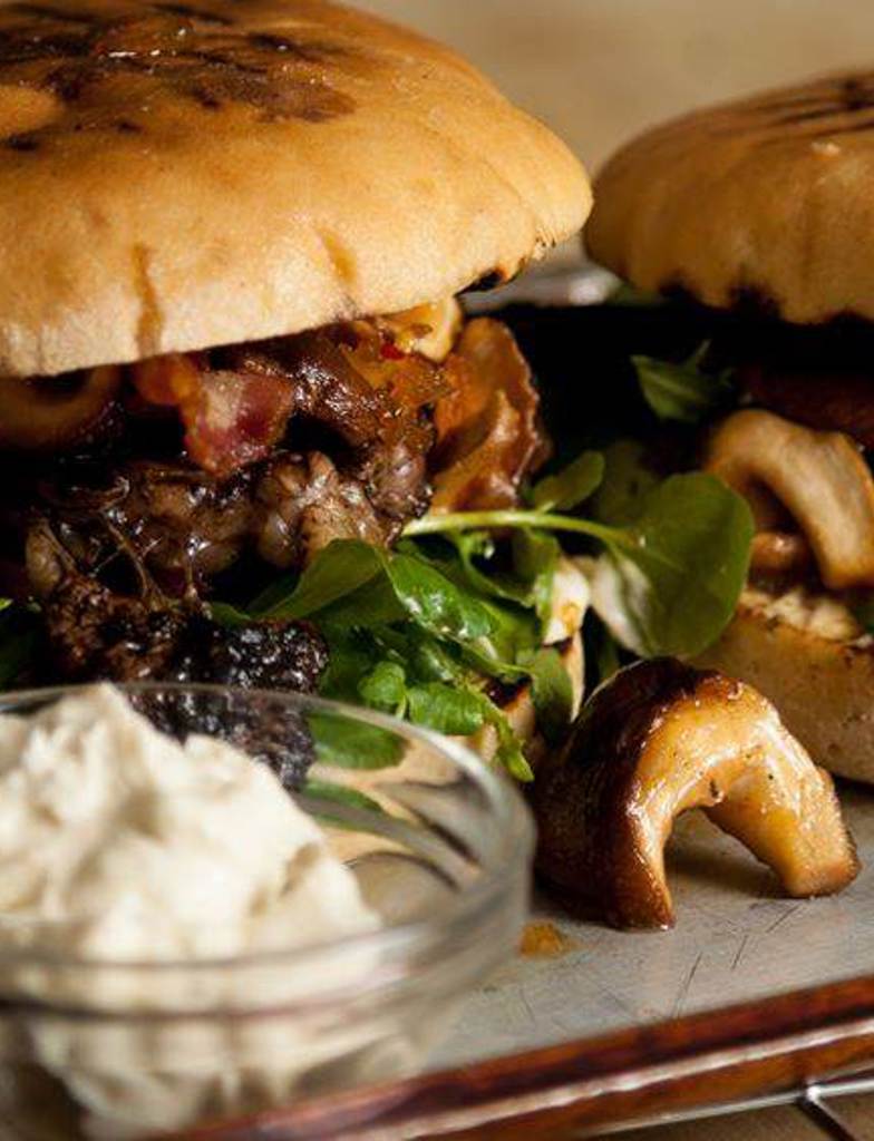 A tray with 2 classic unseeded burger buns filled with steak, caramelized chili mushrooms and onions and a side of onion compote and béarnaise sauce