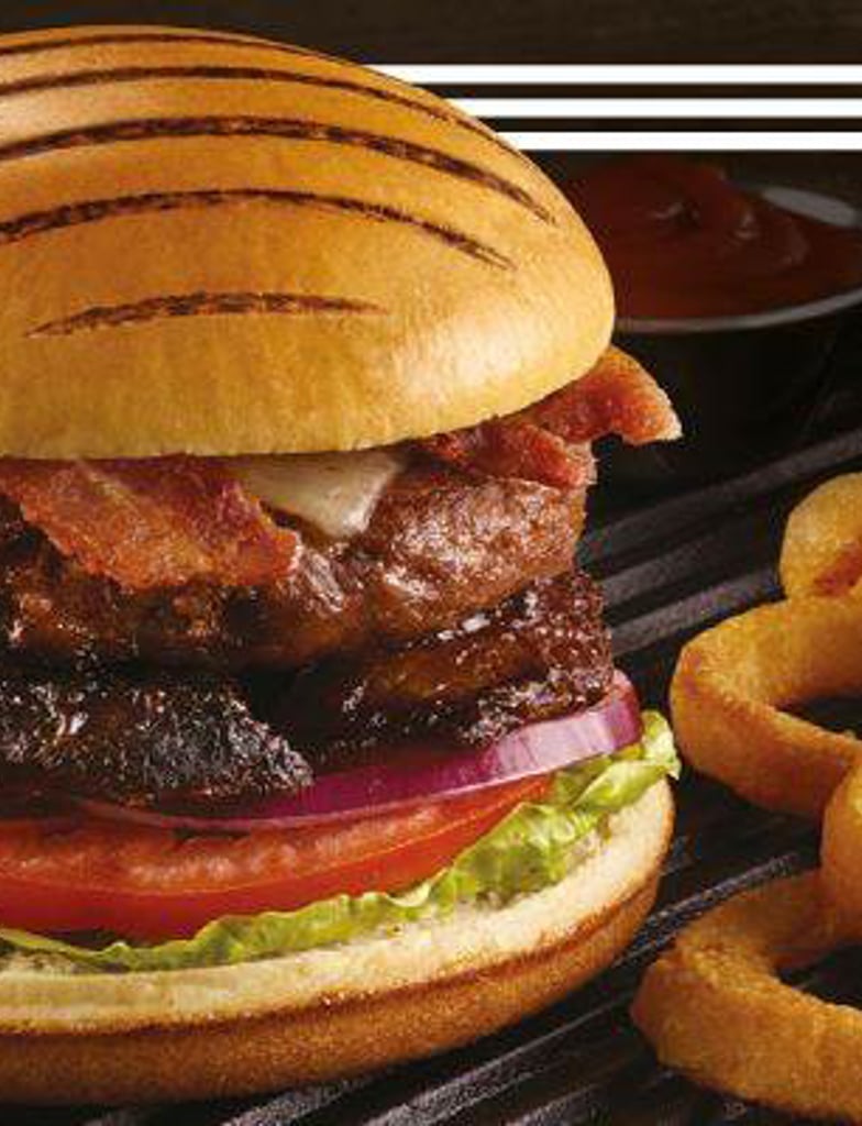The Ultimate BBQ Burger on display with an array of onion rings next to it, the Americana Grill Marked Burger Bun is stacked with lettuce, tomato, red onion, slow cooked brisket, bbq sauce, burger patty, cheese and bacon