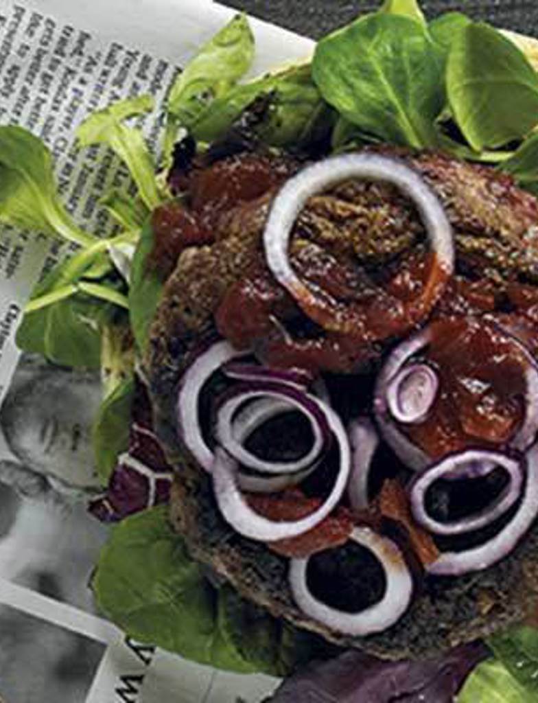 A burger displayed on top of a newspaper, An open face gluten free poppy seed bun with pesto spread on it, the other side has a pile of lettuce, burger patty and tomato relish with red onion on top 